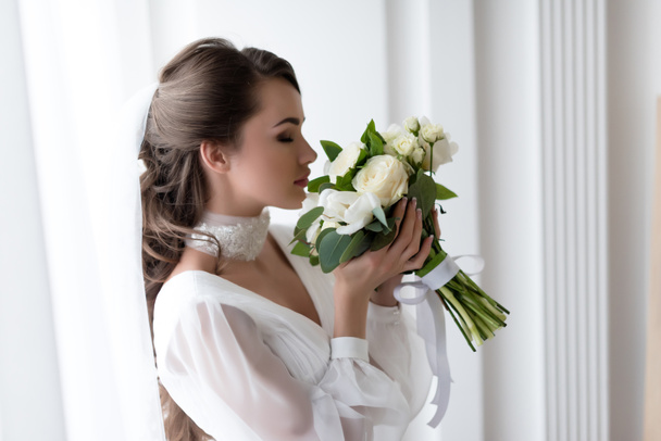 привлекательная невеста в свадебном платье и вуаль, нюхающая белый букет
 - Фото, изображение