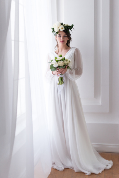 attractive bride in traditional dress and floral wreath holding wedding bouquet  - Φωτογραφία, εικόνα