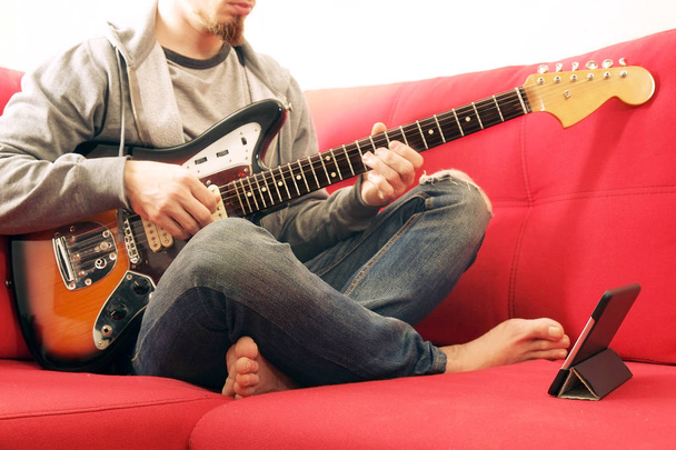 Casually dressed young man with guitar playing songs in the room at home. Online guitar lessons concept. Male guitarist practicing chord grips. - 写真・画像