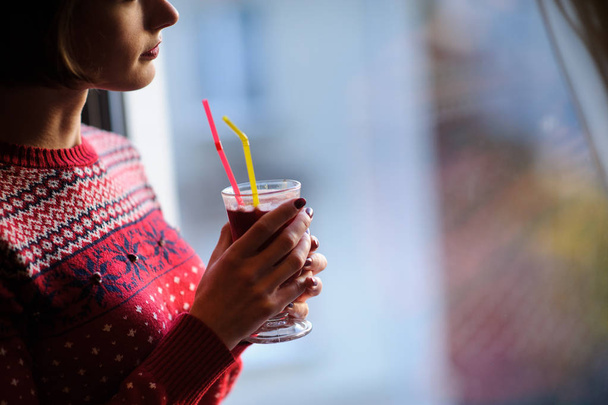 Beautiful woman standing next to a window, wearing red winter clothes and drinking mulled wine - 写真・画像