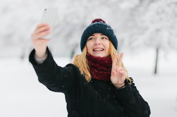 Jonge Kaukasische blond meisje in een zwarte jas en rode gebreide sjaal wandelingen in winter park met smartphone - Foto, afbeelding
