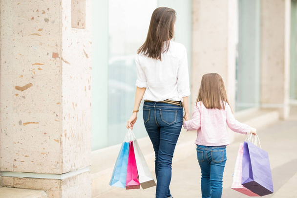 Rückansicht von Mutter und Tochter, die mit Einkaufstaschen an Schaufenstern entlang laufen - Foto, Bild