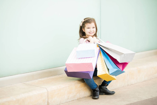 Menina sentada perto da loja com sacos de compras coloridos
 - Foto, Imagem