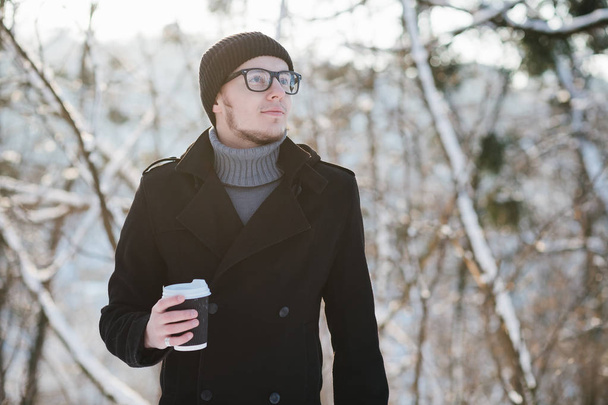 Bir gri kazak ve siyah ceket siyah bir şapka ve gözlük ile genç bir adam portresi kış parkta sıcak kahve içer - Fotoğraf, Görsel