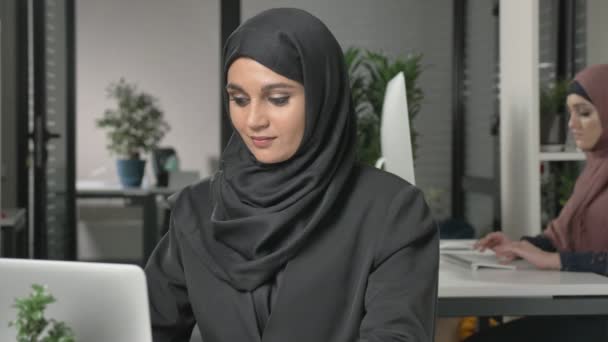 Young beautiful girl in black hijab working at office on computer. Smiling and looking at the camera. Girl in red hijab in the background. Close up 60 fps - Materiaali, video