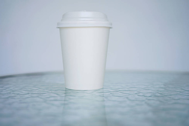 A white paper glass (one-time) for coffee is on the table. Background is blurred, white and blue. Place for the title on the glass. Side view with copy space - Foto, afbeelding