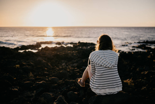 Mädchen, das abends mit Sonnenbrille auf einem Felsen am Meer sitzt. Teneriffa - Foto, Bild