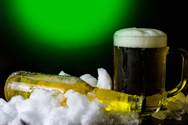 Fles en bier mok in de sneeuw op een groene en zwarte achtergrond - Foto, afbeelding
