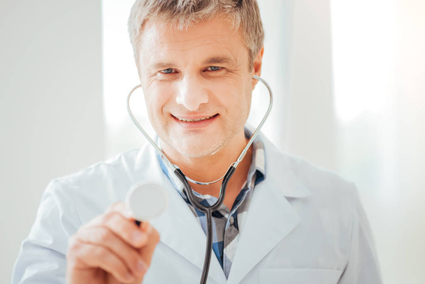 Pleasant satisfied doctor using his statoscope and smiling. - Фото, изображение