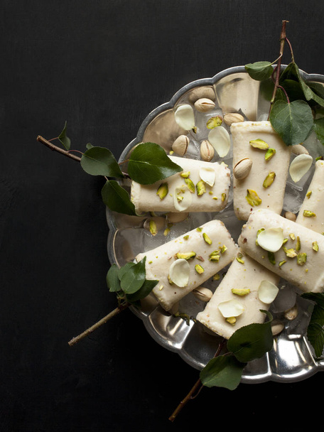 Pista kulfi, indická zmrzlina na servírovací talíř s kostkami ledu, pistácie, okvětních lístků růže, větve stromu se zelenými listy na černém pozadí, pohled shora. Tradiční indická kuchyně Rajasthani. - Fotografie, Obrázek