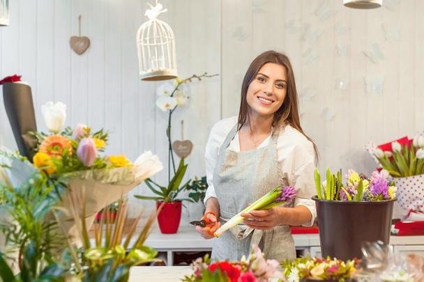 Smiling woman florist small business flower shop owner, at counter holding a scissor cuting hyacinths, making arrangements and smiling at camera. - Photo, Image