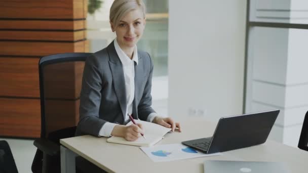 Portrait of Attractive blonde businesswoman sitting at table writing in notebook smiling into camera in modern office - Video