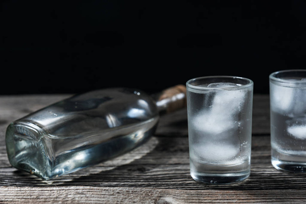 Vodka en vasos de chupito y botella sobre mesa de madera rústica fondo negro
 - Foto, Imagen