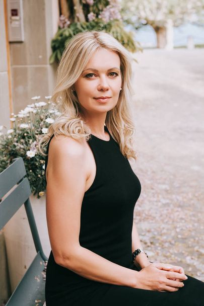 Outdoor portrait of young blond woman wearing black dress, sitting on the bench - Photo, Image