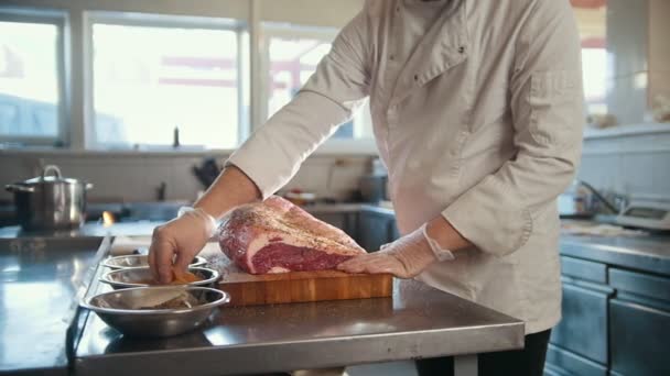 Slager het toevoegen van kruiden in grote stuk van vers rauw vlees, liggend op een houten plank in een commerciële keuken, slow-motion - Video