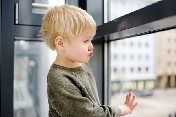 schöner kleiner Junge sitzt am Fenster in der Nähe des Panoramafensters und schaut nach draußen - Foto, Bild