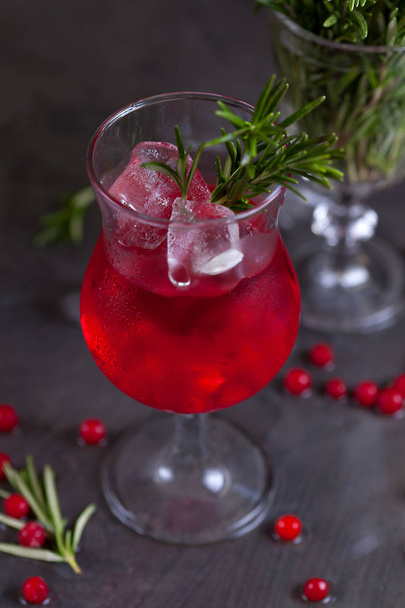 a glass with a cocktail of red color with lime, red berries, rosemary and ice cubes on a background of rosemary on a dark background - Foto, afbeelding