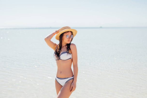 Jonge sexy slank meisje permanent op een strand dragen witte bikini badmode op zee achtergrond - Foto, afbeelding