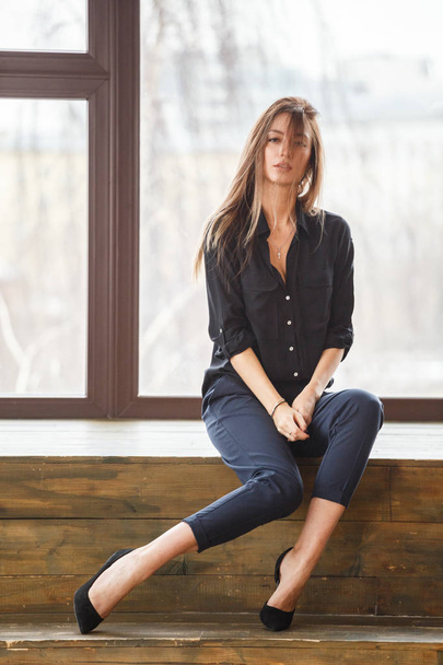 Studio shot of beautiful young woman wearing black casual clothes sitting on wooden floor, lighted windows background - 写真・画像