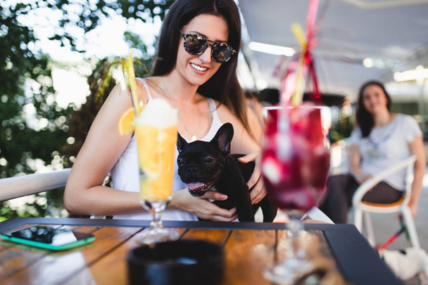 Beautiful young woman with sunglasses sitting in cafe with her adorable French bulldog puppy. People with dogs theme - Fotografie, Obrázek
