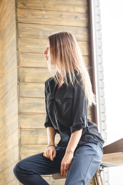 Studio shot di bella giovane donna vestita con abiti casual neri seduta su sedia di legno, sfondo finestra illuminato
 - Foto, immagini