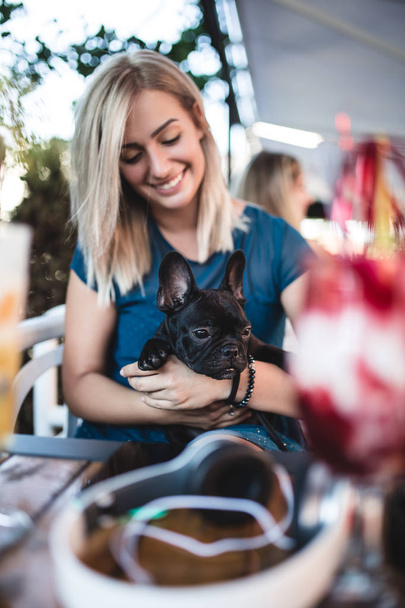 Beautiful young blonde woman enjoying with her french bulldog puppy in cafe bar - 写真・画像