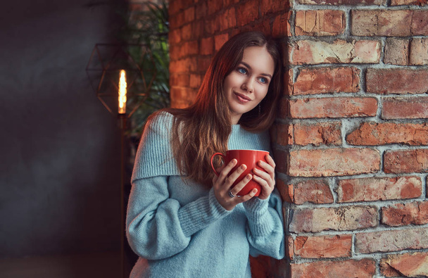 A sensual brunette in a gray sweater drinks coffee leaning against a brick wall in a room with loft interior. - Фото, изображение