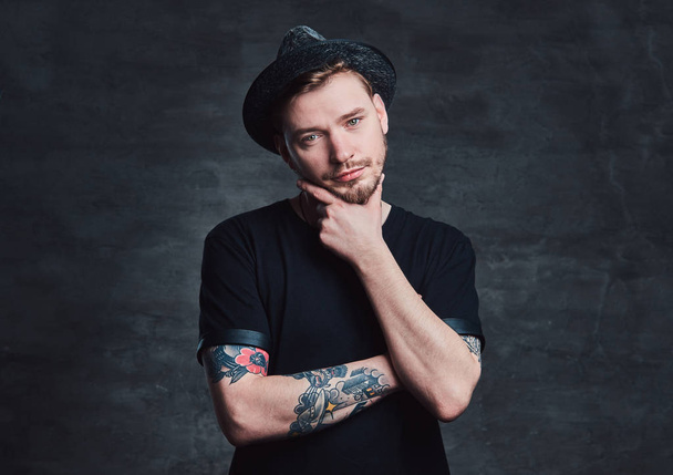 Portrait of handsome bearded hipster with crossed tattooed arms, dressed in a black t-shirt and hat. Isolated on a dark background. - Photo, image