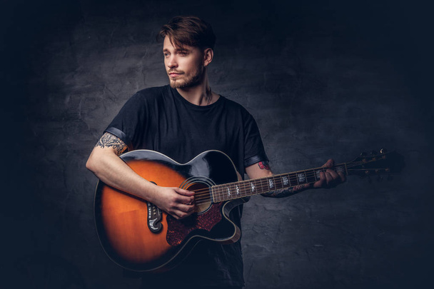 An attractive tattooed guitarist in black t-shirt playing on acoustic guitar, isolated on a dark background. - Valokuva, kuva