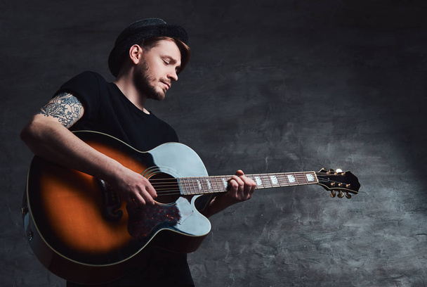 Egy vonzó tetovált gitáros játszik akusztikus gitár, sötét háttér előtt elszigetelt kalapban. - Fotó, kép