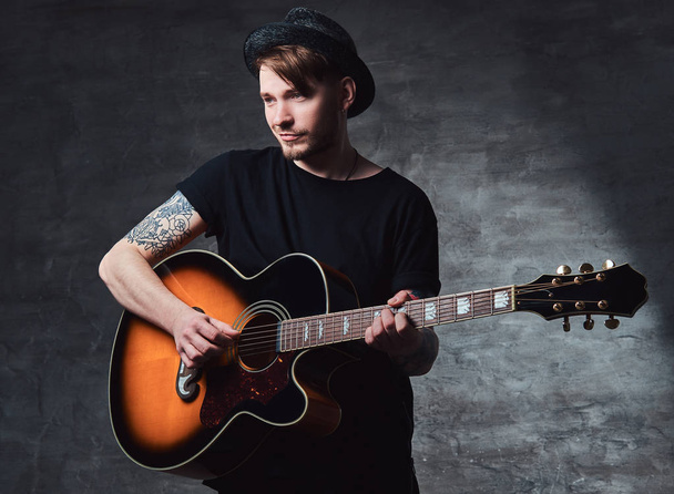 Egy vonzó tetovált gitáros játszik akusztikus gitár, sötét háttér előtt elszigetelt kalapban. - Fotó, kép