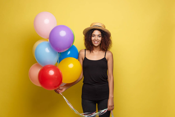 Conceito de celebração - Close up Retrato feliz jovem bela mulher africana em t-shirt preta sorrindo com balão de festa colorido. Estúdio Pastel Amarelo Fundo
 - Foto, Imagem
