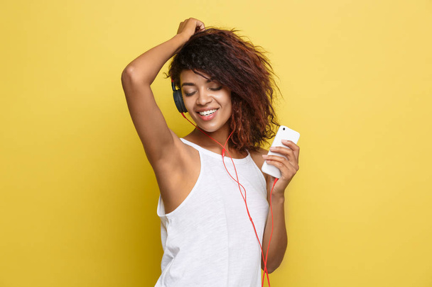 Lifestyle-Konzept - Porträt einer schönen Afroamerikanerin, die freudig Musik auf dem Handy hört. Gelber pastellfarbener Studiohintergrund. Kopierraum - Foto, Bild