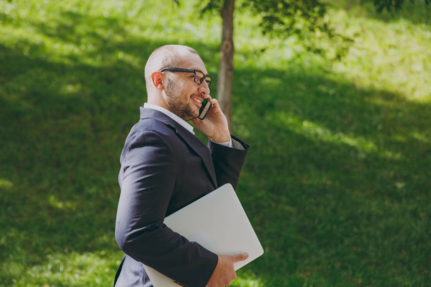 Úspěšný podnikatel smart v bílé košili, klasický oblek, brýle. Muž stojí s pc notebooku, mluvit na mobilním telefonu v městském parku venku na pozadí přírody. Boční pohled, obchodní koncept. - Fotografie, Obrázek