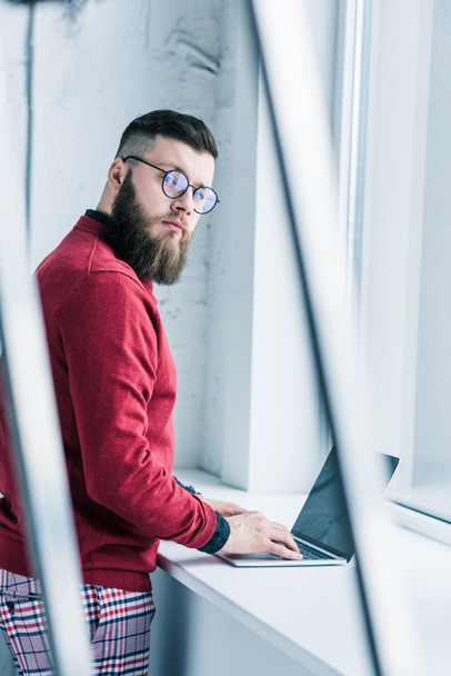 side view of stylish businessman looking at camera while typing on laptop - Photo, Image