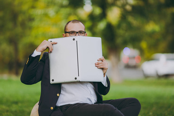 Young successful businessman in white shirt, classic suit, glasses. Man sit on soft pouf, hide behind laptop pc computer in city park on green lawn outdoors on nature. Mobile Office, business concept. - Photo, image