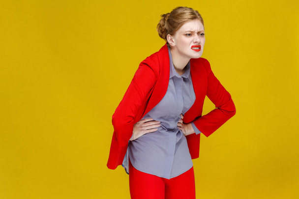 redhead businesswoman in red suit bending in half from pain in belly on yellow background, stomach pain concept  - Fotó, kép