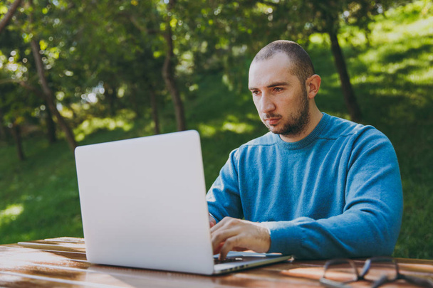 Mladý úspěšný inteligentní muž podnikatel nebo student v ležérní tričko modré brýle sedí u stolu s mobilním telefonem v městském parku přenosném počítači práci venku na zeleném pozadí. Mobilní kancelář koncept. - Fotografie, Obrázek