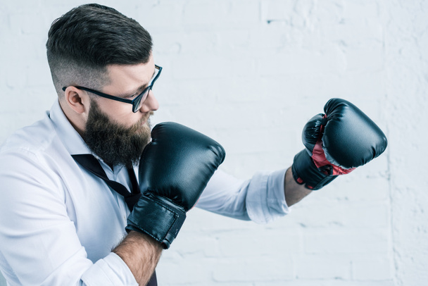side view of bearded businessman in eyeglasses and boxing gloves against white brick wall - Foto, imagen