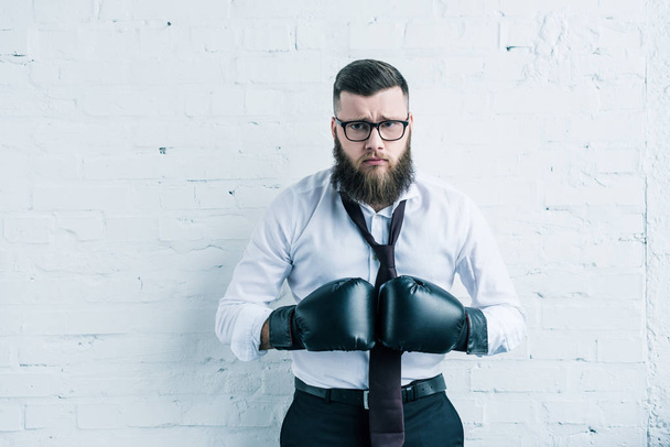 портрет расстроенного бизнесмена в боксёрских перчатках, смотрящего в сторону стены из белого кирпича
 - Фото, изображение