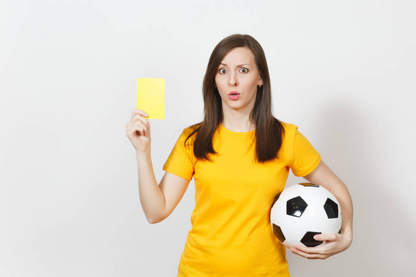 European serious severe young woman, football referee or player in yellow uniform showing yellow card, holding soccer ball isolated on white background. Sport play football, healthy lifestyle concept. - Photo, Image