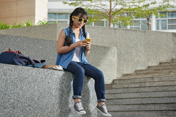 Souriant jeune étudiant assis à l'extérieur et textos sur smartphone
  - Photo, image