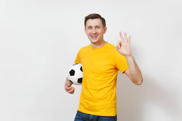 Inspired young European man, fan or player in yellow uniform hold soccer ball show OK gesture cheer favorite football team isolated on white background. Sport play football, healthy lifestyle concept. - Photo, Image