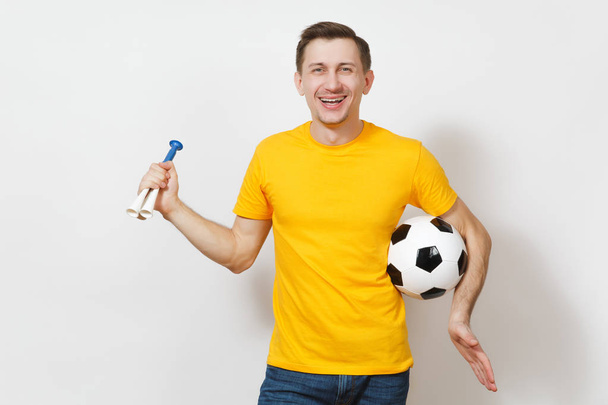 Geïnspireerd jonge leuke vrolijke Europese man, ventilator of speler in geel uniform houdt voetbal, pipe, cheer favoriete voetbalteam geïsoleerd op een witte achtergrond. Sport, voetballen, levensstijl concept. - Foto, afbeelding