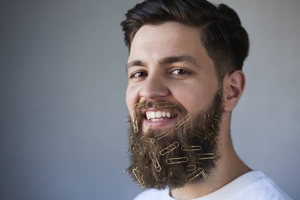 Close-up van jonge man gezicht met paperclips op baard, grijze studio achtergrond - Foto, afbeelding