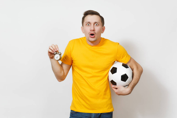 Inspired young cheerful European man, fan or player in yellow uniform hold soccer ball, old alarm clock, cheer favorite team isolated on white background. Sport, play football, lifestyle concept. - Photo, Image