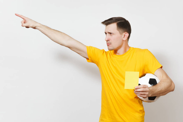 Σοβαρή σοβαρή Ευρωπαϊκή νεαρός, διαιτητής ποδοσφαίρου ή παίκτη σε κίτρινο ομοιόμορφη δείχνει κίτρινη κάρτα, κρατώντας την μπάλα ποδοσφαίρου που απομονώνονται σε λευκό φόντο. Αθλητισμός, ποδόσφαιρο παιχνίδι, έννοια του υγιεινού τρόπου ζωής. - Φωτογραφία, εικόνα