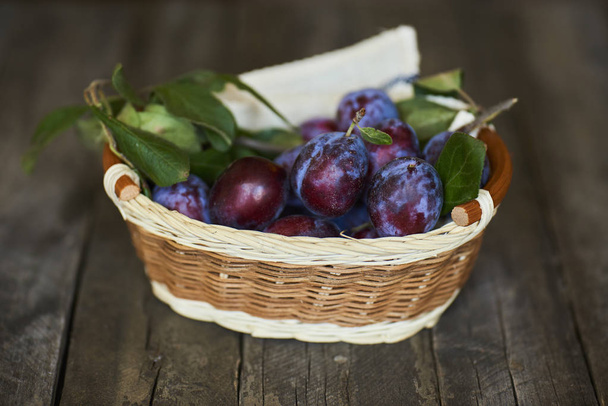 Fresh plums with green leaves in wicker basket on wooden table - Photo, Image