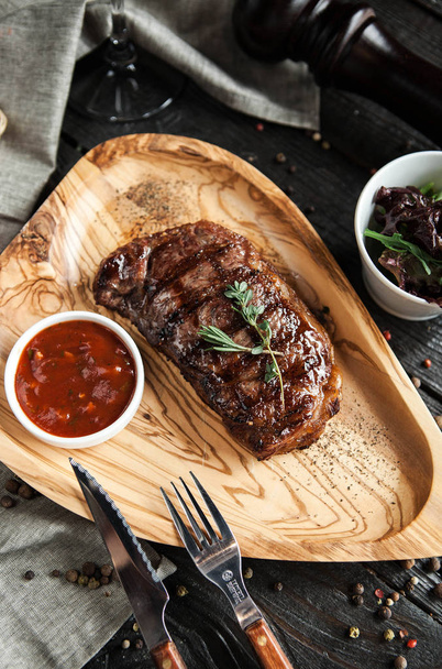 belle portion de steak de viande sur une assiette en bois avec salade, sauce tomate et un verre de vin rouge sur un fond sombre en bois
 - Photo, image