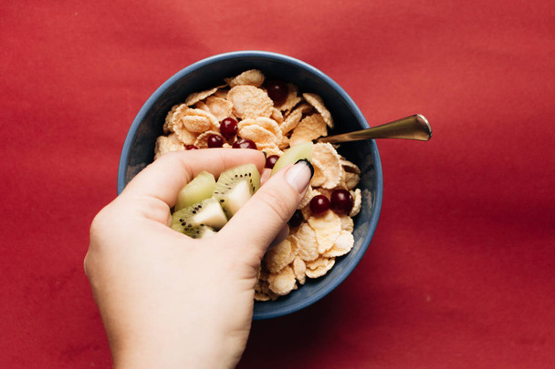 female hand adding kiwi to bowl with crispy cornflakes and cranberries on red background,  healthy breakfast - Zdjęcie, obraz